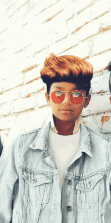 a boy wearing sunglasses and a denim jacket stands in front of a brick wall