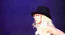 a blonde woman wearing a black hat and glasses waves her hand