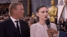 a woman in a white dress is being interviewed by a man in a suit and tie at the oscars .