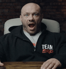 a bald man wearing a black team warp sweatshirt sits at a desk