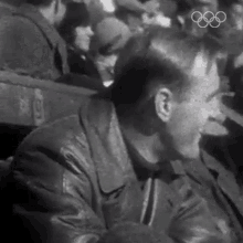 a man in a leather jacket is sitting in a crowd of people with the olympic rings in the background