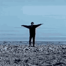 a man is standing on a rocky beach with his arms outstretched in front of the ocean .