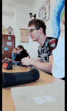 a boy wearing glasses sits at a desk in a classroom with a pencil case that says adidas on it