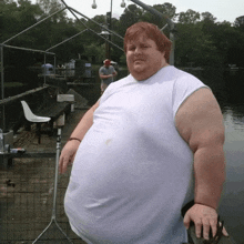 a very fat man in a white shirt stands in front of a body of water