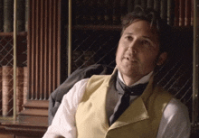 a man wearing a yellow vest and tie is sitting in front of a bookshelf