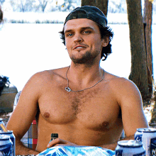 a shirtless man wearing a hat and a necklace is sitting in front of a table full of cans of bud light