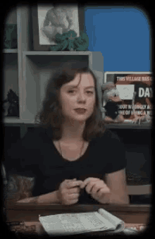 a woman in a black shirt is sitting at a desk in front of a sign that says " this village has a day "