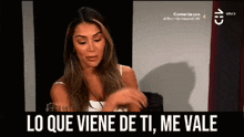 a woman in a white tank top is sitting in front of a sign that says lo que viene de ti me vale