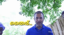 a man in a blue shirt is standing in front of a tree and talking to someone .