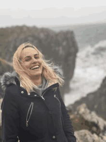 a woman wearing a black jacket with a fur hood is smiling in front of the ocean