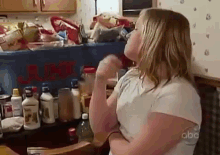 a young girl is standing in front of a messy kitchen counter with bottles of cleaning supplies on it .