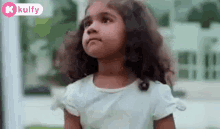 a little girl with curly hair is standing in front of a building and looking up at the sky .