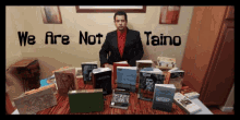 a man sits at a table surrounded by books with the words " we are not taino " behind him