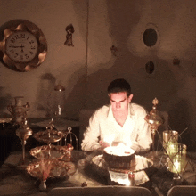 a man is sitting at a table with a cake and candles in front of a wall clock that shows the time as 10:11