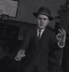 a black and white photo of a man in a suit and hat making an ok sign .