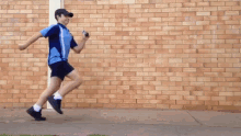 a boy in a blue shirt and black shorts is running on a sidewalk in front of a brick wall