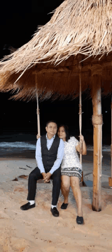 a man and woman sit on a swing under a thatched roof on the beach