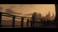 a group of people are walking along a pier with a bridge in the background that says ' brooklyn ' on it