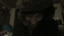 a person wearing a furry hat looks at the camera in a dark room