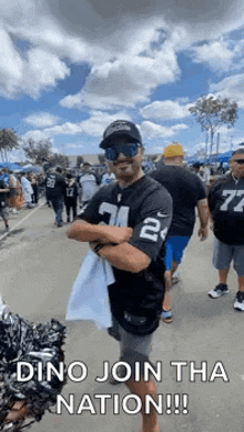 a man wearing sunglasses and a raiders jersey is standing in a crowd of people with his arms crossed .