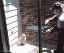 a man in a baseball cap is standing next to a cat in front of a brick wall that says gifakne