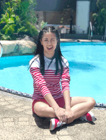 a girl in a red and white striped shirt sits in front of a swimming pool