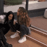 two women are squatting down on a set of stairs . one of the women is wearing white sneakers .