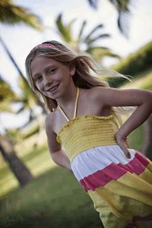 a young girl wearing a yellow white and pink dress stands in front of palm trees