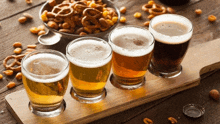 four glasses of beer are on a wooden tray with pretzels in the background