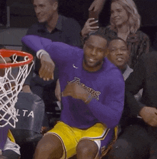 a man in a purple shirt and yellow shorts is sitting in front of a basketball hoop giving a thumbs up .