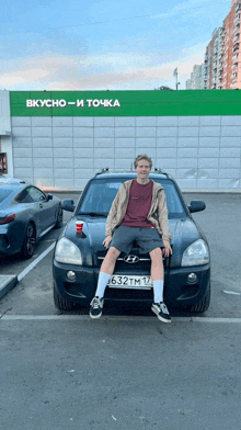 a man sits on the hood of a hyundai car in front of a building that says вкусно-и точка