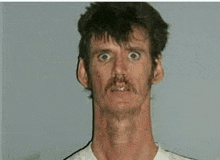 a man with a very long neck and a mustache is looking at the camera .