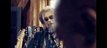 a man wearing sunglasses is singing into a microphone in front of a bunch of guitars .