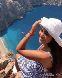 a woman wearing a white hat is sitting on a cliff overlooking a body of water with boats in it