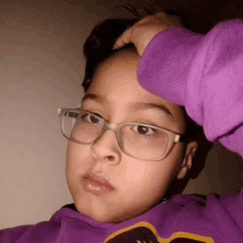 a young boy wearing glasses and a purple hoodie is touching his head .