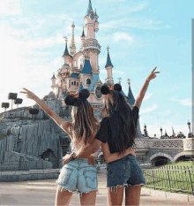 two women wearing mickey mouse ears are posing in front of a castle .