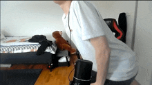 a man in a white shirt is squatting in a room with a teddy bear on a bed .