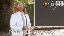 a man in a lab coat and tie stands in front of a blackboard that says " natural process "