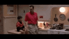 a man in a red shirt is standing in a kitchen next to a boy sitting on a counter .