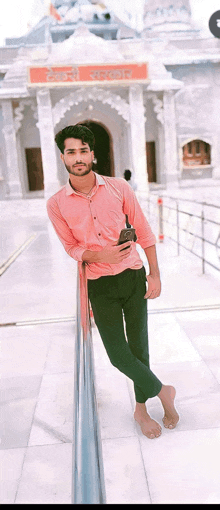 a man in a pink shirt and green pants leans against a railing holding a cell phone