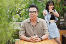 a man wearing glasses sits at a table with his hands folded and a woman behind him pointing