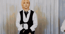 a young man with blonde hair is standing in front of a white curtain wearing a white shirt and a black vest .