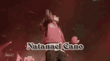 a man in a red jacket stands on a stage with the name natalias cano above him