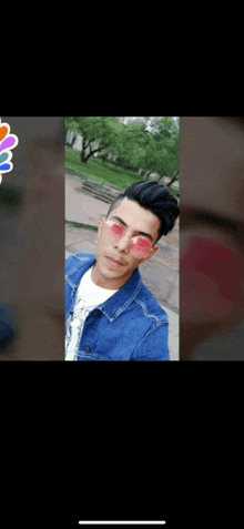 a young man wearing a denim jacket and pink heart shaped sunglasses is taking a selfie .