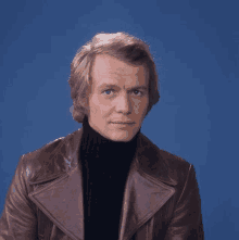 a man wearing a brown leather jacket and a black turtleneck is sitting in front of a blue background .