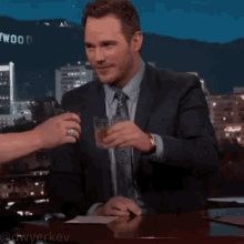 a man in a suit and tie is toasting with a glass of whiskey in front of a hollywood sign