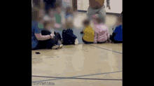 a group of children are sitting on the floor in a gym with a man standing in the background .