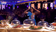 a woman in a blue dress stands in front of a table full of food with the words strictly come dancing on the bottom right