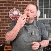 a man in an apron is eating a sandwich in front of a brick wall with a clock in the background