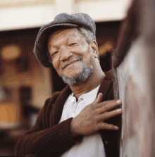 an older man with a beard and hat is leaning against a wall .